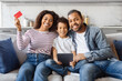 A joyful African American family is gathered on the sofa in a cozy, well-lit living room, using digital tablet. Mother happily displays a credit card, suggesting online shopping or financial planning