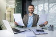 © Liubomir - Smiling businessman sitting at a desk in a modern office, holding papers, with a laptop and calculator nearby. Portrays a productive and positive work environment.