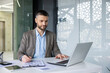 © Liubomir - A focused business man reviewing documents while working on his laptop in a modern office setting, with natural light backlighting the scene.