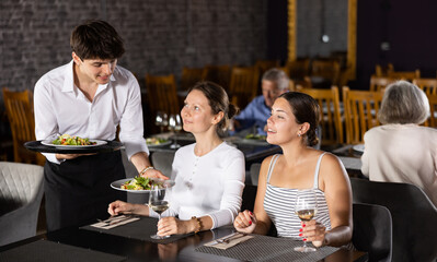 Wall Mural - Polite young waiter serving fresh vegetable salads to positive female guests visiting restaurant for dinner, sitting at table with glasses of wine ..