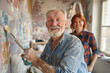 A cheerful senior male artist, with a full beard and twinkling eyes, confidently applies paint to a canvas. His female assistant, with red hair and a plaid shirt, stands behind him. 