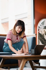 Wall Mural - Young woman is engrossed in writing notes next to her open laptop, showcasing productivity and multitasking.