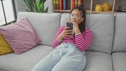 Wall Mural - A young hispanic woman relaxes on a sofa with her phone, surrounded by colorful pillows in a cozy living room.