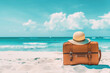 A tan suitcase with a straw hat on top of it is sitting on the sand near the ocean