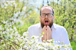 Man allergic suffering from seasonal allergy at spring in blossoming garden at springtime. Young bearded man with glasses sneezing in front of blooming tree. Spring allergy concept