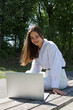 © MM - A woman in a white shirt and white pants sits on a park bench with a laptop in her hands.