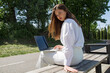 © MM - A woman in a white shirt and white pants sits on a park bench with a laptop in her hands.