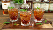 Three glasses of mint julep on a wooden board, with ice cubes and fresh mint leaves, bourbon bottles blurred in the background.