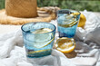Summer refreshing lemonade drink or non alcoholic cocktail with lemon slices in blue drinking glasses on white blanket in the garden. Fresh healthy cold lemon beverage. Water with lemon. Summer mood.