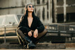 Fashion street. Sexy fashion woman sitting outdoor. Dangerous areas. Fashionable brunette in black jacket bandana and sunglasses in urban style. Lifestyle of female hipster. Casual dress style.