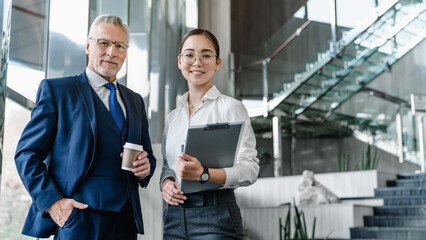 Wall Mural - Portrait of mature smiling business man and business woman standing posing in office. Two diverse colleagues, group team of confident professional business people.