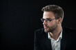 Portrait of young confident guy in eyeglasses over black background with look away. Side view shot of bearded freelancer boss ceo teacher manager in formal clothes, suit. Profile picture