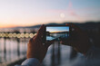 Cropped image of man taking photo in evening dusk of nature environment using modern smartphone camera, back view of hipster guy holding mobile phone making picture of sea water via app in twilight