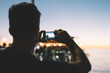 Back view of man taking picture of evening scenery near sea using modern mobile phone camera, male tourist making photo in night twilight via contemporary application for photographing in dark