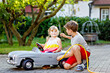 Two happy children playing with big old toy car in summer garden, outdoors. Kid boy refuel car with little toddler girl