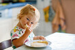 Happy little toddler girl eating delicious pancakes sitting in the kitchen. Cute child tasting different food