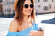 Young beautiful smiling woman in trendy summer clothes. Carefree female posing in street at sunny day. Positive model holds smartphone, looks at cellphone screen, uses mobile apps, in hat