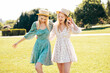 Two young beautiful smiling hipster woman in trendy summer dress. Carefree women in the street in hats. Positive models at sunset. Cheerful and happy. Walking in grass, enjoy summertime days