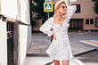 Young beautiful smiling blond woman in trendy summer white dress. Sexy carefree woman posing in street  at sunset. Positive model outdoors at sunny day. Cheerful and happy. In hat, sunglasses