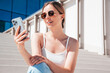 Young beautiful smiling hipster woman . Carefree woman posing in the street. Positive model holds mobile phone, looks at cellphone screen, uses smartphone apps. Sites at stairs