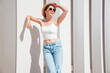 Beautiful smiling model in sunglasses. Female dressed in summer hipster white T-shirt and jeans. Posing near white wall in the street. Funny and positive woman having fun outdoors, in hat, sunglasses