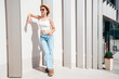 Beautiful smiling model in sunglasses. Female dressed in summer hipster white T-shirt and jeans. Posing near white wall in the street. Funny and positive woman having fun outdoors, in hat, sunglasses