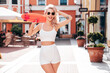 Young beautiful smiling female in summer white cycling shorts and tank top clothes. Sexy carefree woman posing in street. Positive model . Cheerful and happy. Holds penny skateboard, in sunglasses