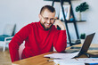 Cheerful hipster guy laughing at funny content in social networks sitting at desktop with laptop computer,smiling prosperous male freelancer happy about completed online project successfully