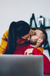 Young marriage kissing while man sitting at desktop with laptop computer for remote job and online business, woman hugging her husband with love feeling romantic and love being together at home