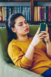Pensive brunette woman reading book online via gadget on smartphone sitting in armchair near bookcase, serious hipster girl checking mail on mobile phone connected to wireless internet in library