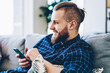 Cheerful hipster guy enjoying internet connection at home for chatting in social networks via smartphone, positive young man satisfied with resting at living room checking notification on cellular
