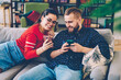 Cheerful romantic couple watching funny video from social networks on mobile phone resting on sofa, positive hipster girl showing common photos on smartphone to her boyfriend laughing together