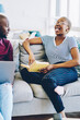 Emotional dark skinned woman making funny face laughing at story from book sitting next to boyfriend using laptop computer, cheerful african american couple spending free time at living room