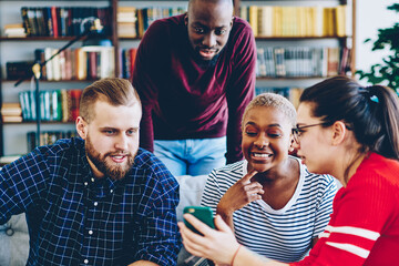 Wall Mural - Cheerful group of male and female friends watching online video on smartphone sitting at couch at living room, hipster girl shoving photos on mobile phone to multiracial colleagues  having fun