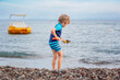 Adorable little blond kid boy building pebble stone castle on beach. Funny child playing with bucket and shovel. Vacations, summer, travel concept. Toddler enjoying summer vacations on sea.