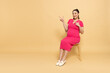 Happy Asian Pregnant woman sitting on wood chair and pointing to copy space isolated on brown background, Motherhood and Pregnancy Concept, Full body composition