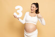 Portrait of Happy Asian pregnant woman standing and holding 3 number or three isolated on brown background
