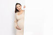 Asian pregnant woman is standing behind the white blank banner and pointing to empty copy space advertisement board on red background, Looking at camera