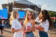 © artiemedvedev - Group of joyful women with cool summery drinks at beach music festival. They dance under clear sky, stage with live performance in background. Girls in sunglasses enjoy fest vibes, sunlit ambiance.