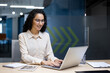 © Liubomir - Happy and smiling hispanic businesswoman typing on laptop, office worker with curly hair and glasses happy with achievement results, at work inside office building