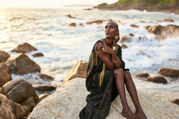 Wall Mural - Non-binary ethnic fashion model in dress, brass jewelry sits on rocks by ocean. Trans sexual black person with rings, nose-ring, bracelets, earrings in posh clothes poses in tropical seaside location.