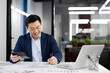 A professional Asian businessman reviews financial documents while smiling in a brightly lit modern office environment, portraying efficiency and contentment.