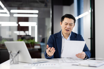 Wall Mural - A businessman looks perplexed while examining documents at his work desk in a modern office with bright, linear lighting.