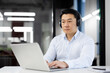 An Asian businessman works focused on his laptop in a modern office setting, wearing headphones and a blue shirt.