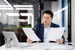 Focused Asian businessman in a blue suit reviews paperwork while working at his desk in a well-lit modern office. Concept of business analysis and professional diligence.