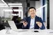 A cheerful Asian businessman holds a credit card and smartphone, sitting at a modern office desk. He appears confident and ready for a business transaction.