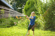 Funny little boy playing with garden hose in sunny backyard. Preschooler child having fun with spray of water. Summer vacation in the village for kids.