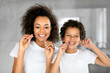 © Prostock-studio - A cheerful African American mother and her young son are standing together in a well-lit, contemporary bathroom, wearing white t-shirts while they both floss their teeth