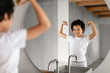 © Prostock-studio - African American young child is standing in front of a bathroom mirror, proudly flexing their muscles and exhibiting a joyful smile. The bathroom has a modern design with visible wooden beams