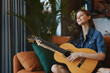 Young woman playing acoustic guitar on a couch in living room at home interior